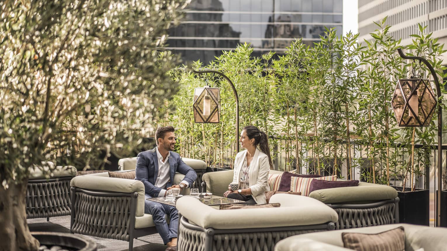 Man in suit and women in white blazer seated on outdoor patio with tree surround