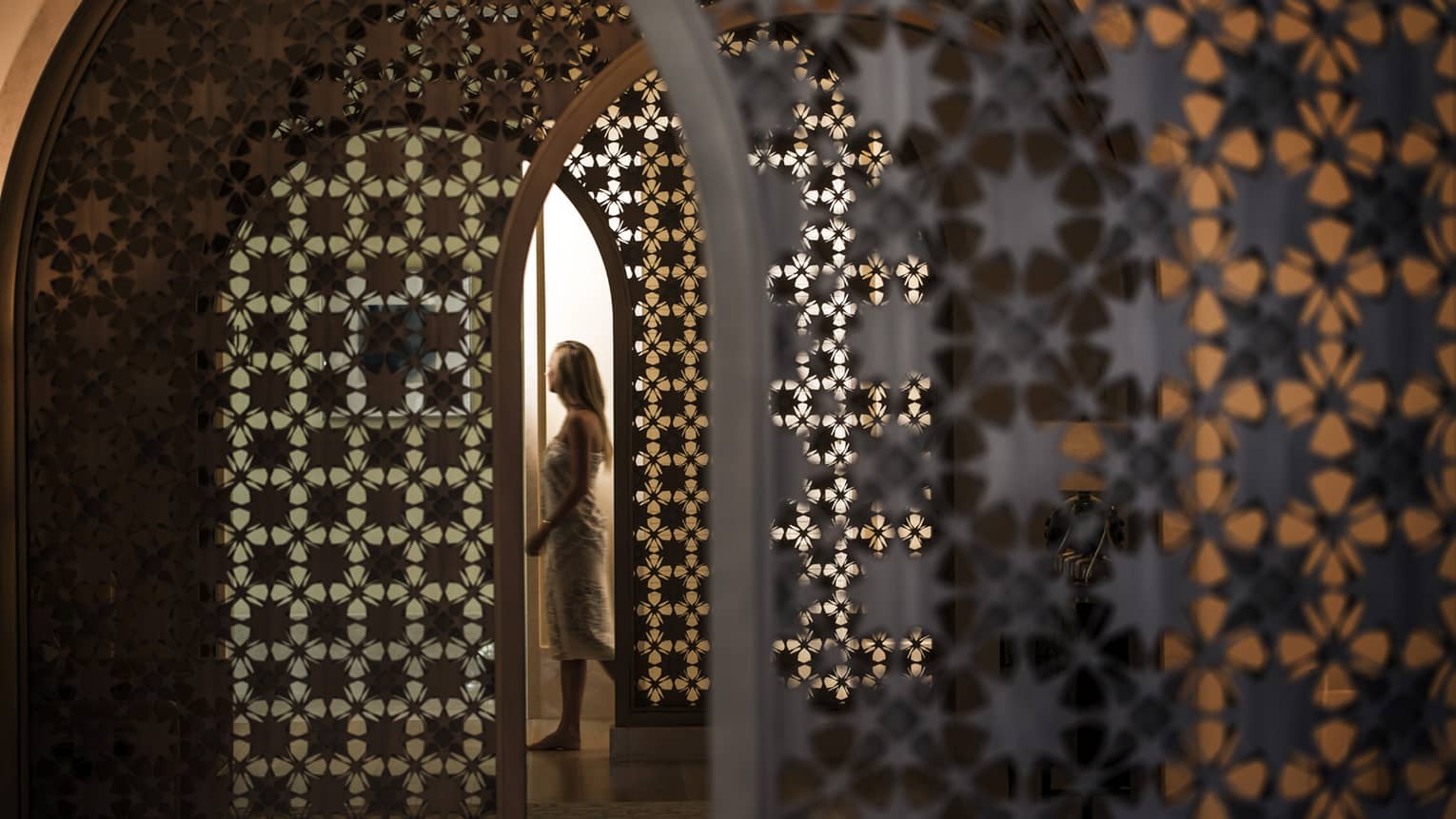 Woman in white wrap walks through dimly-lit decorative entranceway in hall
