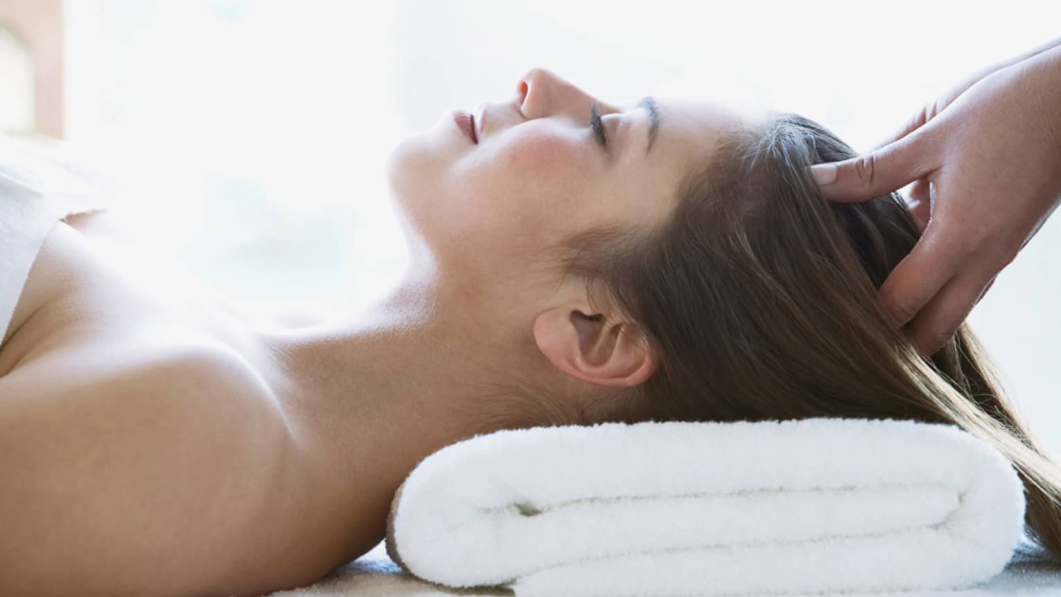 Woman lays on back with head on folded white towel, closes eyes as hands massage her scalp
