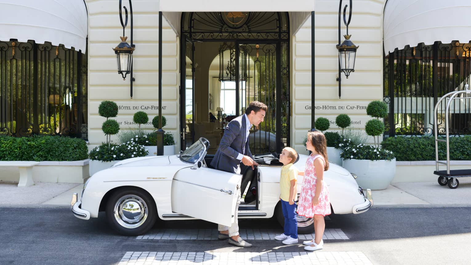 Concierge talking to boy in yellow shirt, girl in pink sundress beside white vintage convertible, in front of hotel