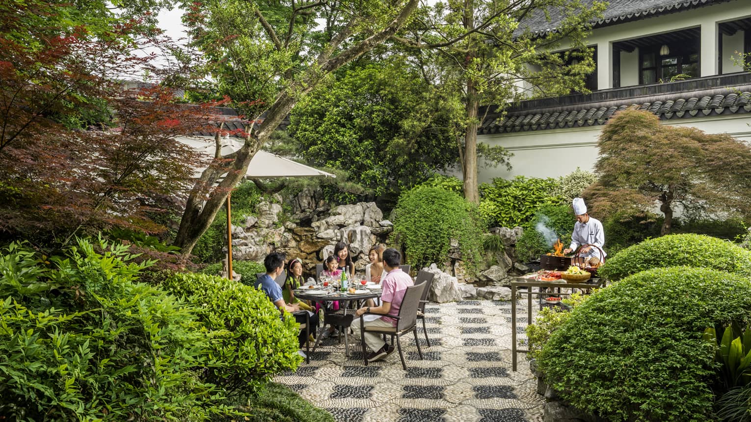 Family dines at round patio table by chef at barbecue in garden
