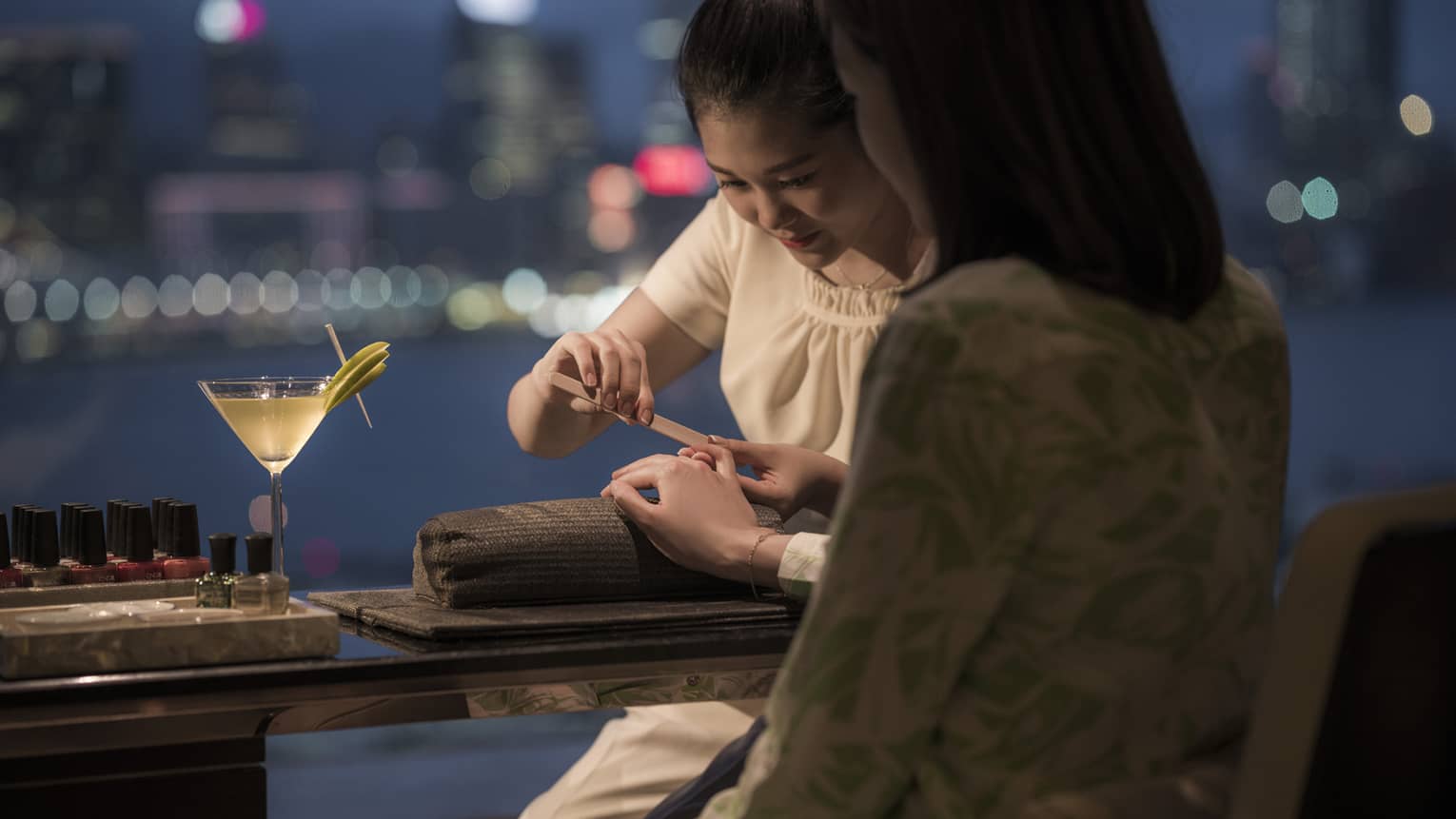 Back of woman getting manicure in spa at night in front of window overlooking harbour, martini on table