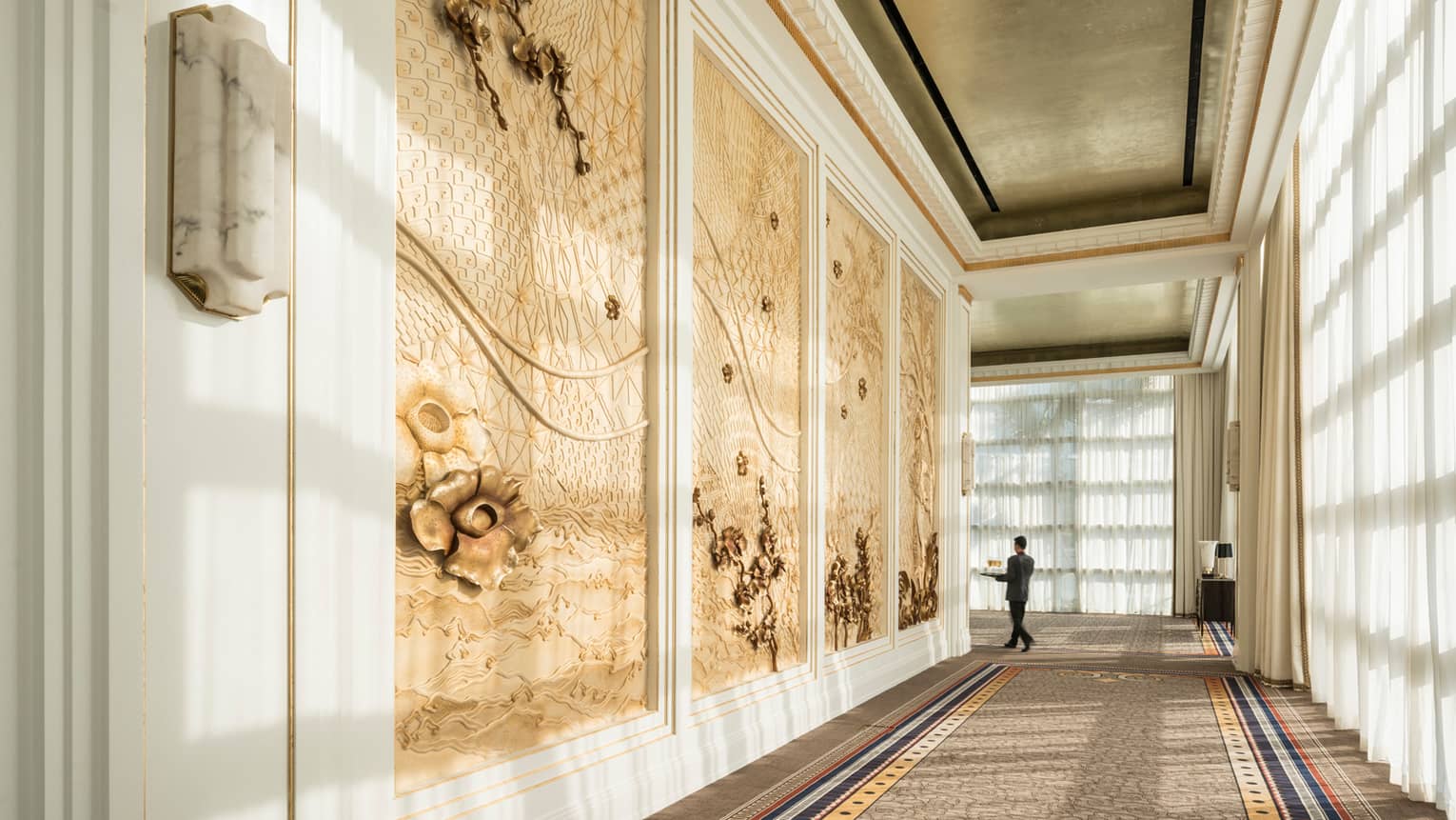 Hotel staff carries tray at end of long hallway past decorative paneled walls