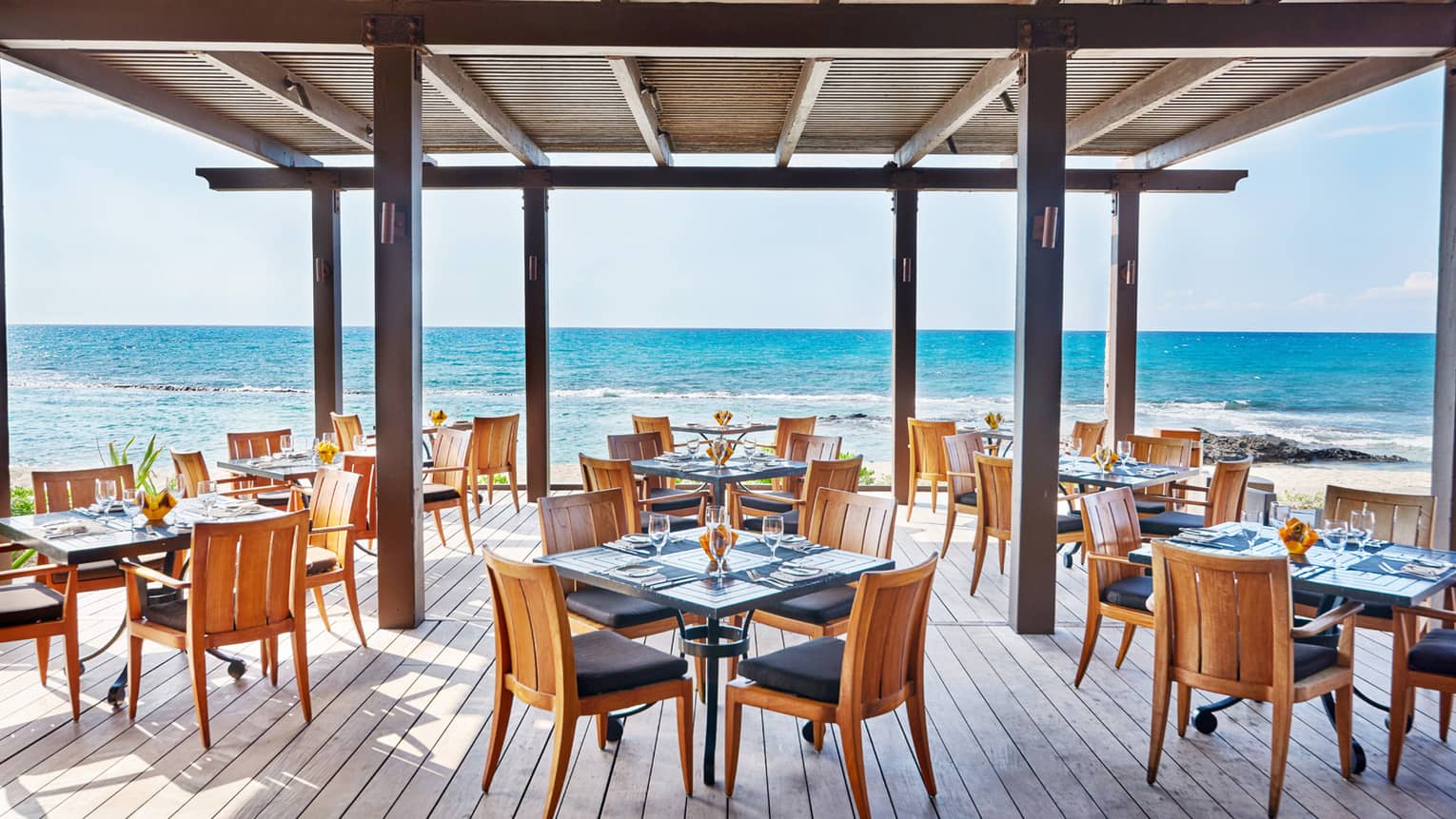 The Ulu Ocean Grill and Sushi Lounge is ready for guests with square tables set with menus and clear glassware, and overlooking the ocean from the covered wooden terrace.
