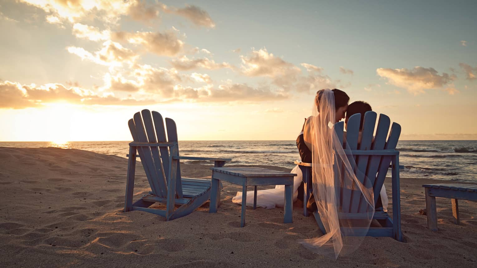 Back of bride and groom sharing chair on sand beach at sunset