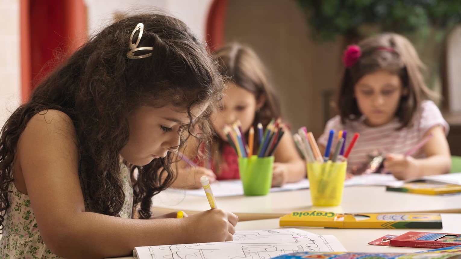 Three girls sitting at a table concentrate on their coloured pencil drawings.