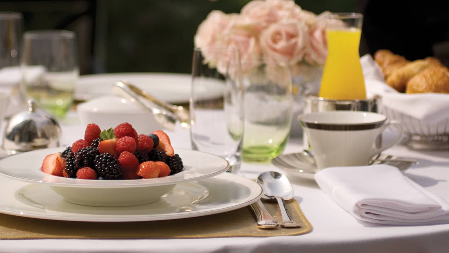White bowl filled with fresh strawberries, blueberries and blackberries on table with glasses, orange juice, pink roses