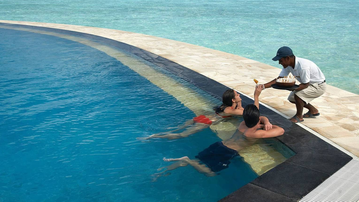 A man and woman swim up to the side of the poolside bar, a smiling server passes a refreshment from tray