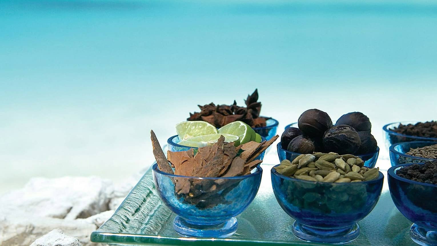 Assortment of spices and herbs in blue glass bowls on a tray with a view of the turquoise sea in the background
