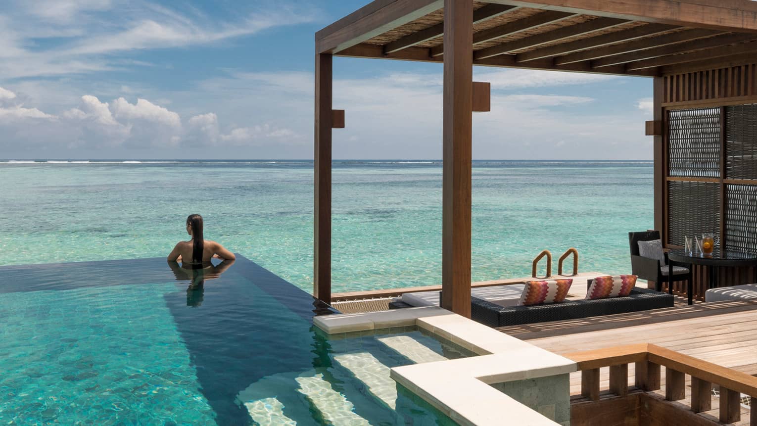 A woman relaxes in the shade of an infinity pool adjacent to a wooden cabana.