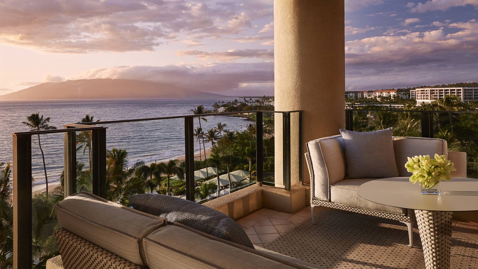Balcony with cushioned seating and table overlooking beach palm trees and ocean at sunset