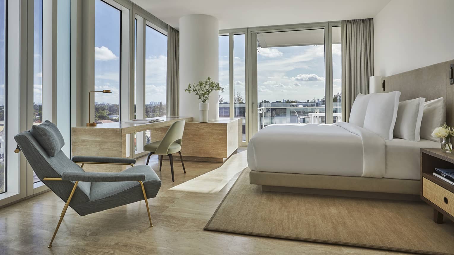 Luxury hotel room with a king bed, floor-to-ceiling windows, a desk, an armchair and a balcony overlooking the city under a blue sky