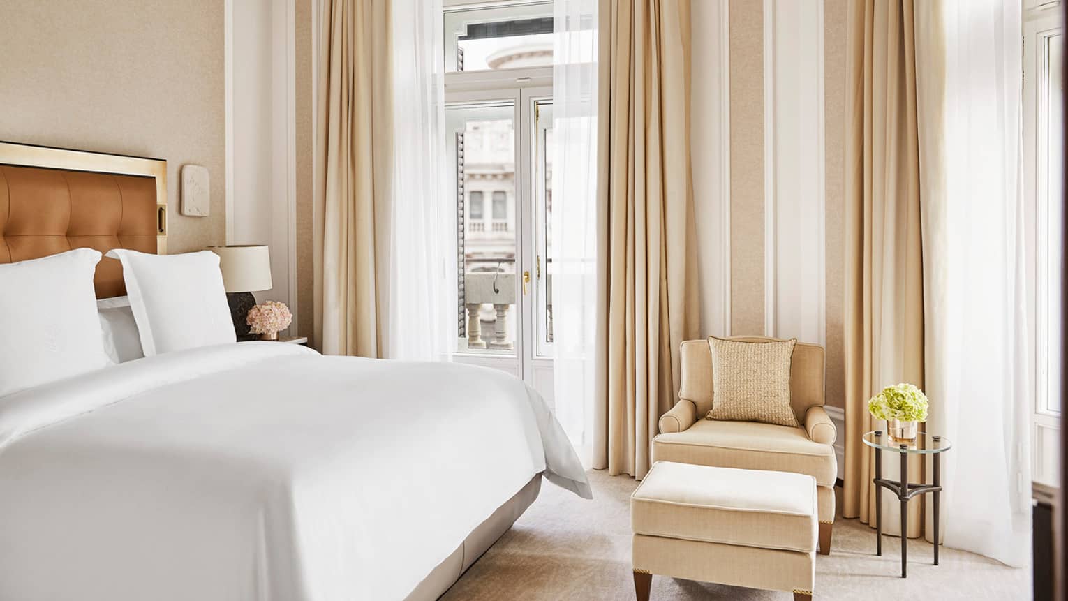 Bedroom with king bed, white linens, beige arm chair and ottoman, large windows and curtains