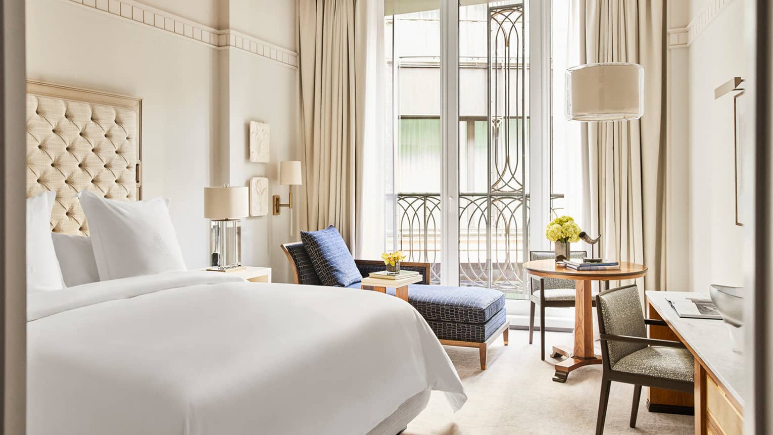 Hotel room with king bed, white linens, off-white walls and floor, dark blue chaise, large windows