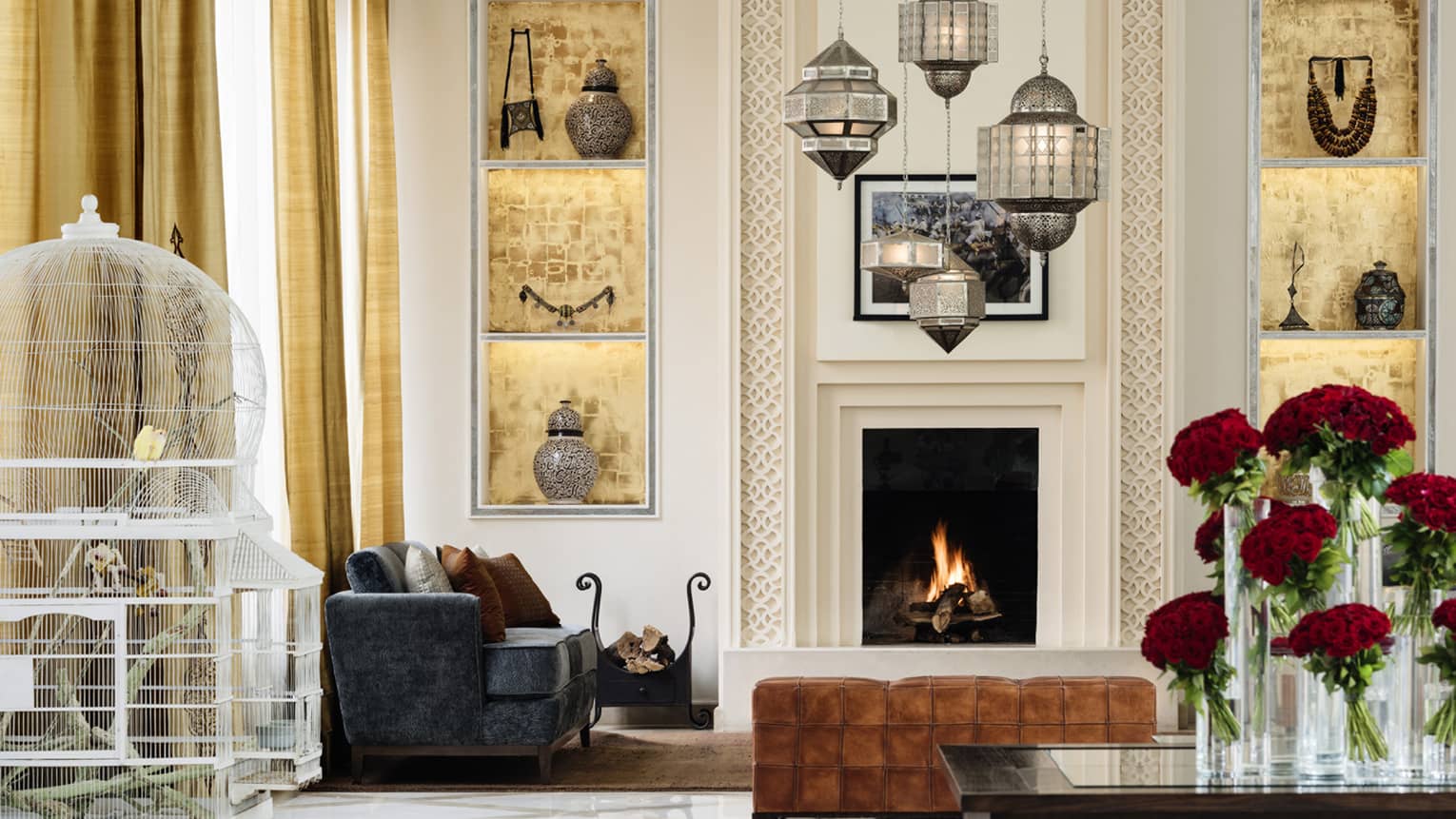 Birds in white cage, red roses in vase in modern hotel suite with sofas, chandeliers by fireplace