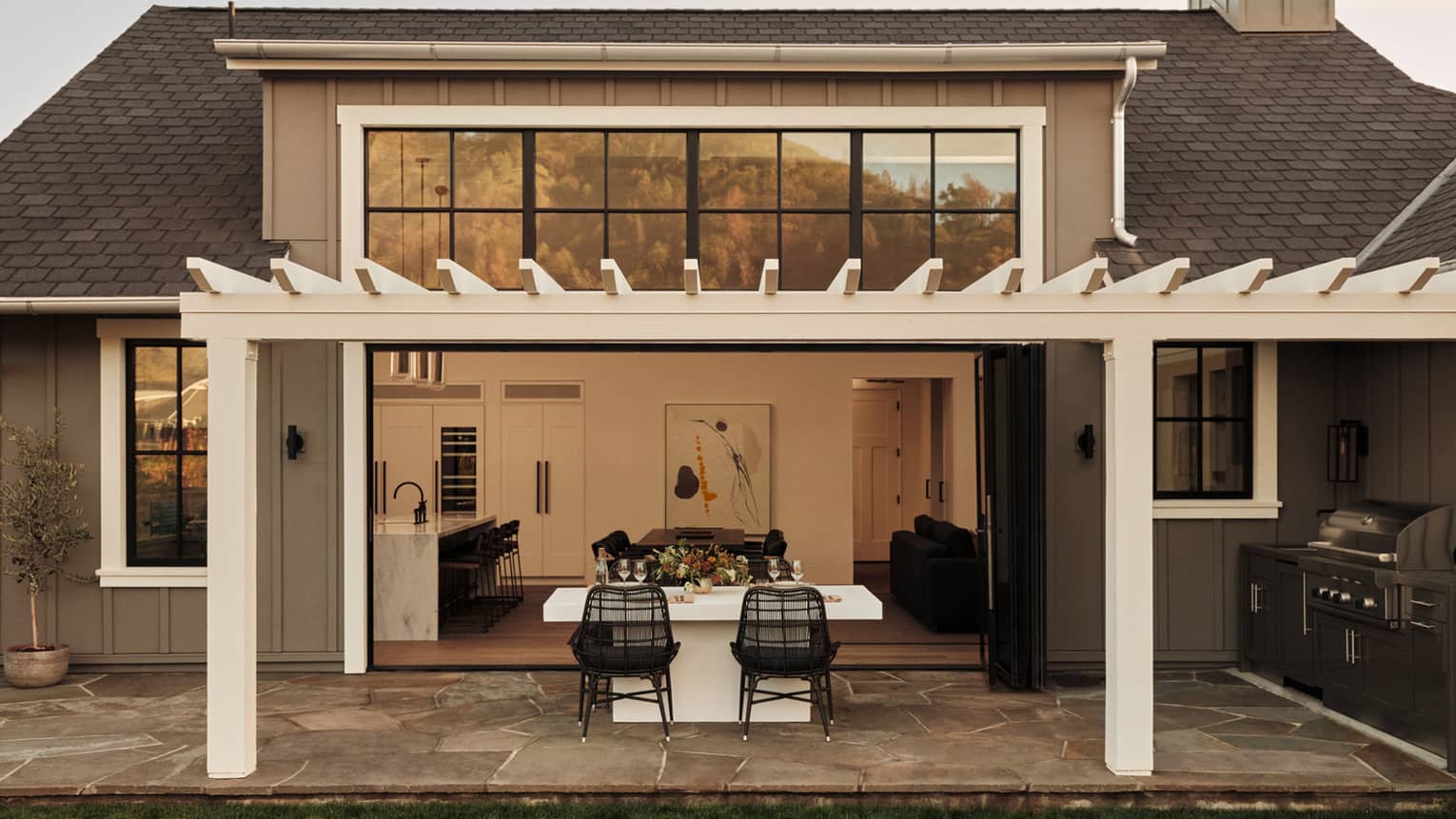 Outdoor dining area of a residential villa with pergola, white table and black wicker chairs, opening to a kitchen and living room through large folding doors