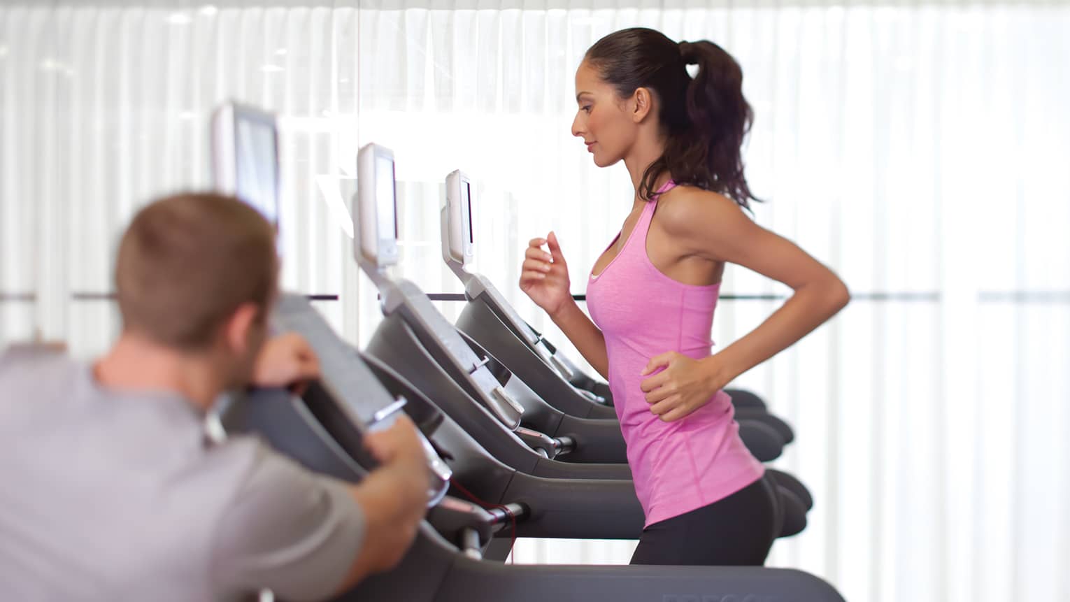 Woman in pink sleeveless shirt and ponytail runs on treadmill as personal trainer watches