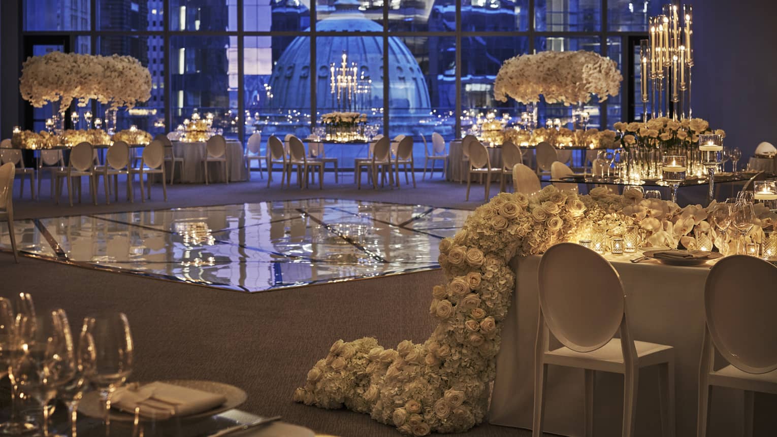 The dance floor in the ballroom is surrounded by white tables decorated with extravagant white flowers and set with white and clear dishes and glasses and overlooks downtown philadelphia through floor to ceiling windows