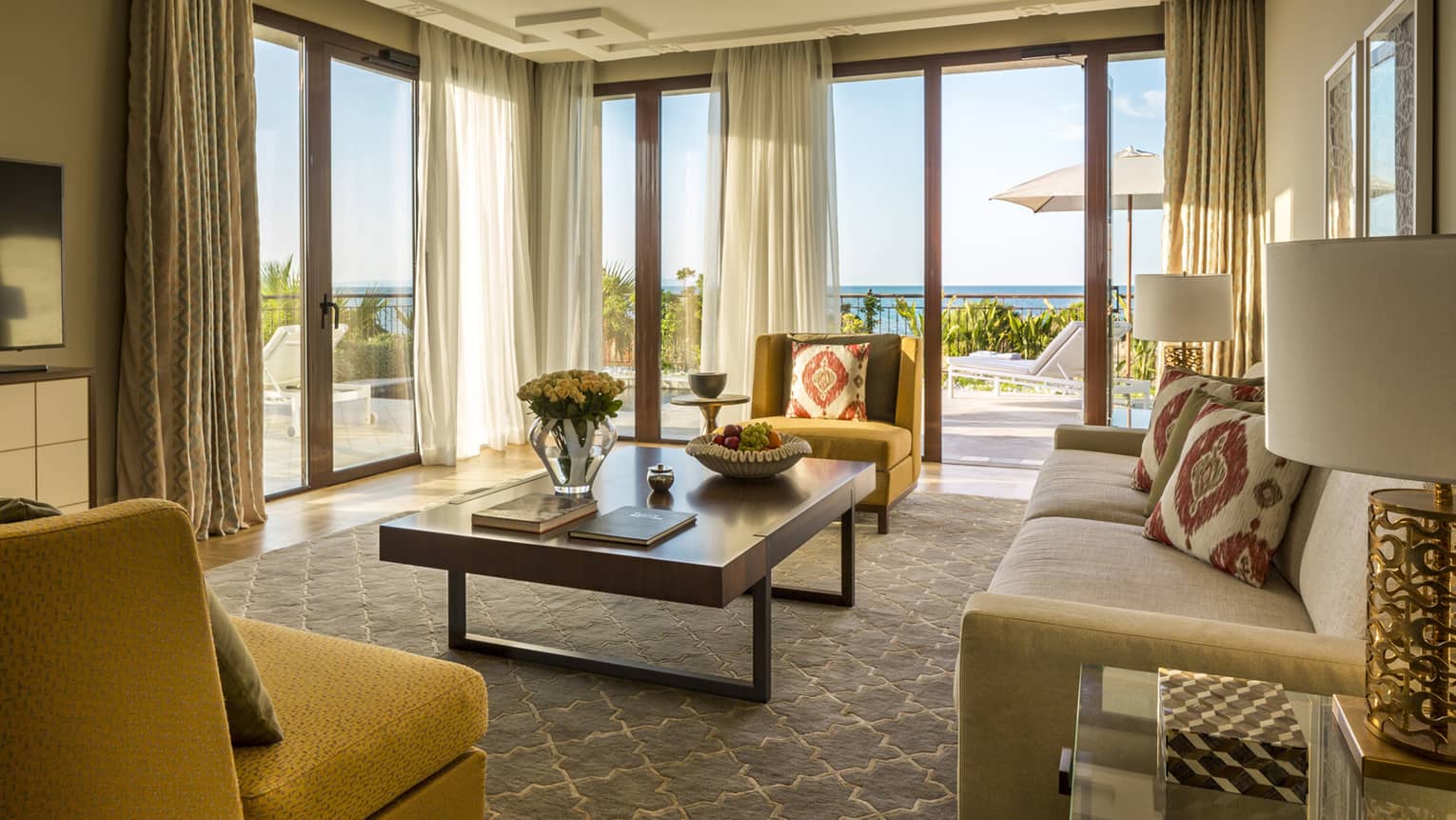 Living room with large rectangular table, beige sofa, mustard-yellow chairs, walls of windows looking out to sea
