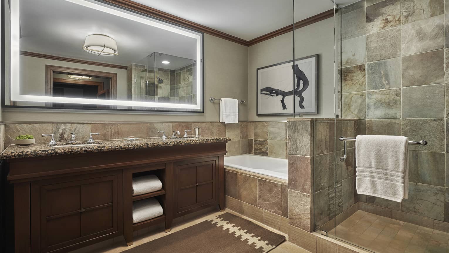 A tiled bathroom, with a double vanity, large lighted mirror and a shower and tub separated by a glass partition