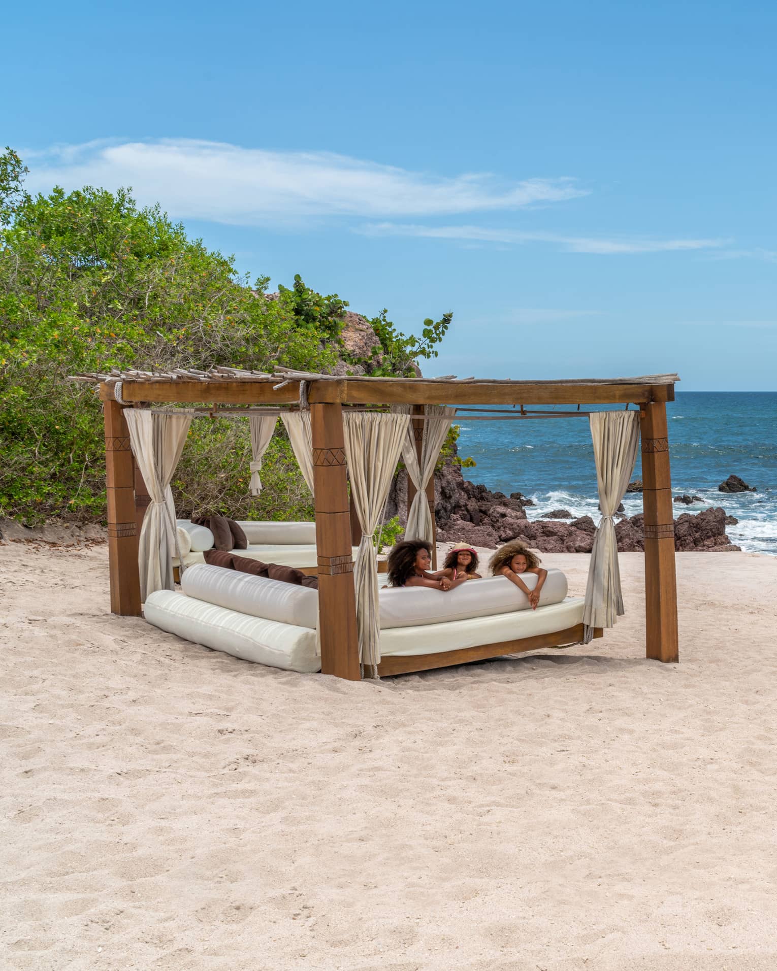 A cabana with three children laying on a plush seat underneath it on the beach, the ocean and plants are in the background.