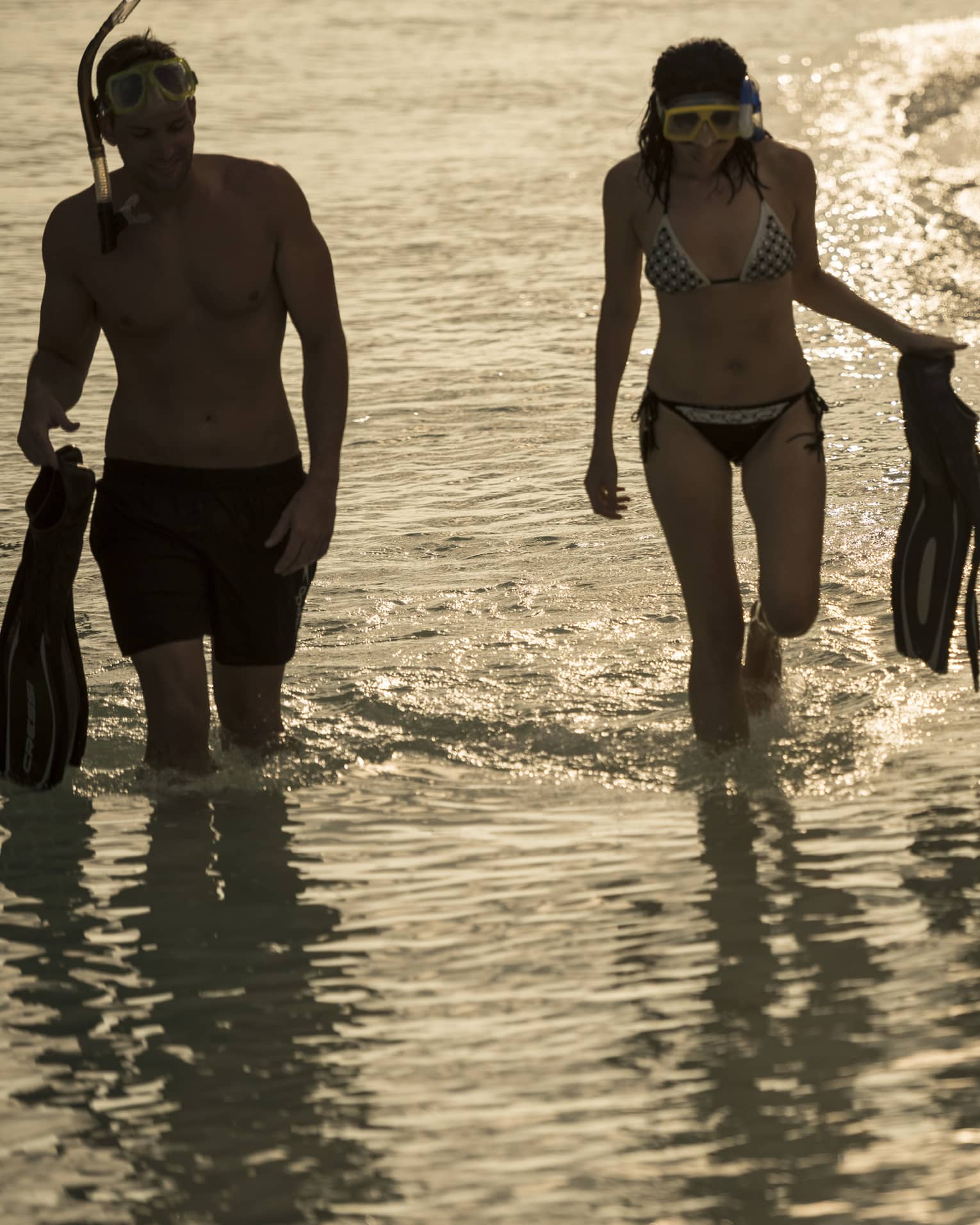 Two people in swimsuits and snorkel gear hold fins as they walk along the water's edge, sun reflecting off gentle waves.