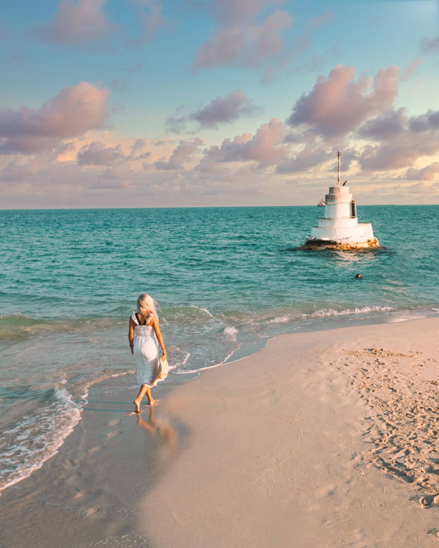 A woman walks along the shoreline towards a stone structure in the sea.