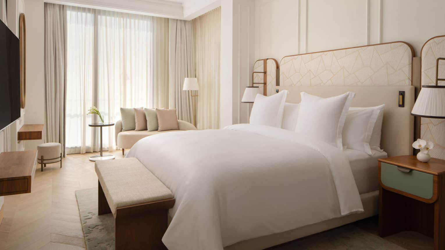 Spacious bedroom with a king bed, padded bench, soft lighting and a seating area near large curtained windows.