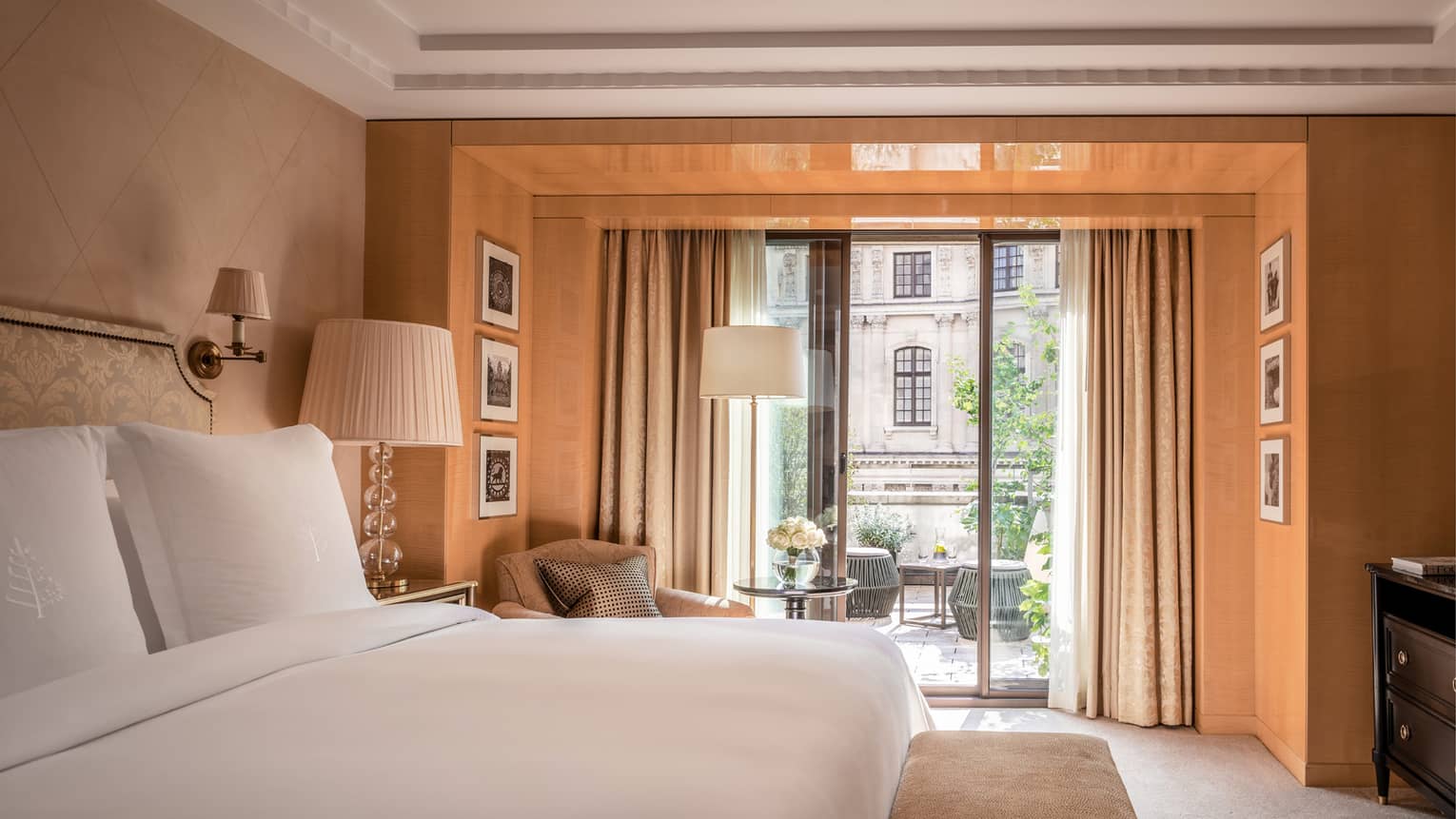 The Grand One Bedroom Suite at Four Seasons London Park Lane in Mayfair features an elegantly designed bedroom with a large, plush bed covered in pristine white linens. The room is bathed in warm light from floor-to-ceiling windows that open to a charming private balcony overlooking a historic London street. The decor includes tasteful gold and cream curtains, stylish bedside lamps, and a small seating area with a modern glass table and a comfortable chair. Framed black and white photographs on the walls add a touch of sophistication to the peaceful and luxurious atmosphere.