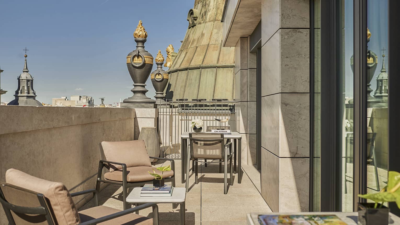 Sunny luxury hotel terrace with outdoor seating, small tables, decorative urns and views of historic architecture and spires