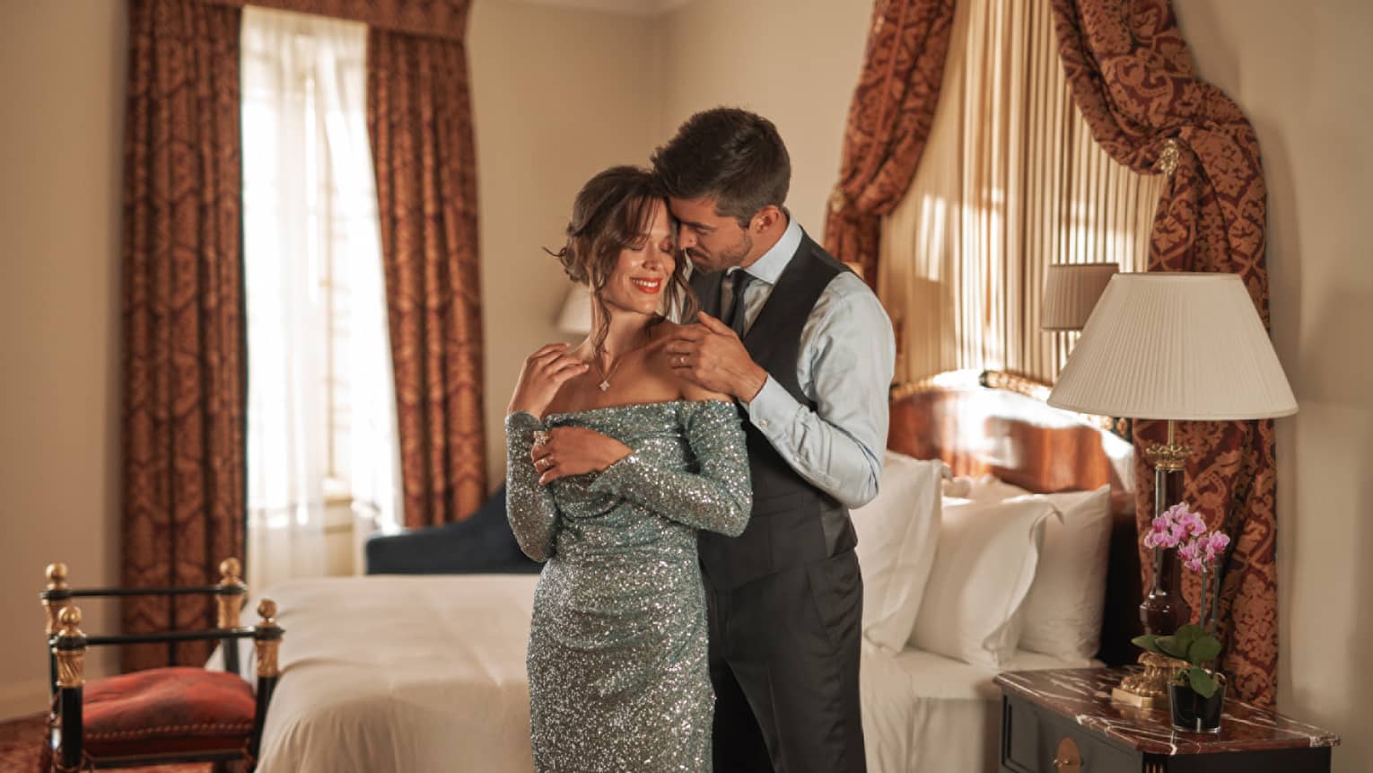 Couple in black tie attire embrace in front of bed in guest room