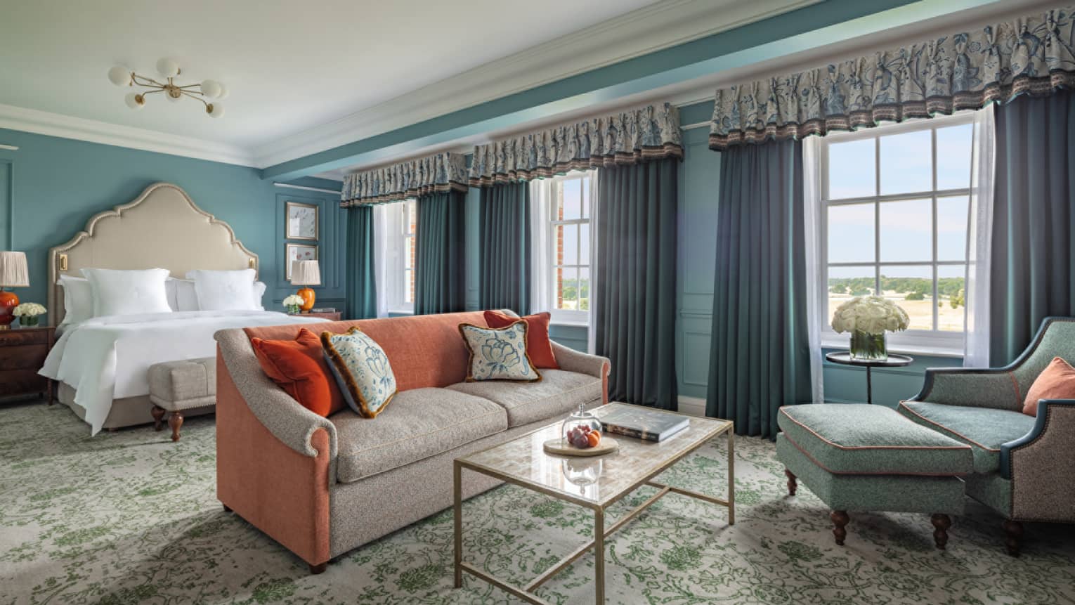 Guest room with Queen bed, cream and red couch, coffee table, blue accent chair, trio of windows with blue drapes