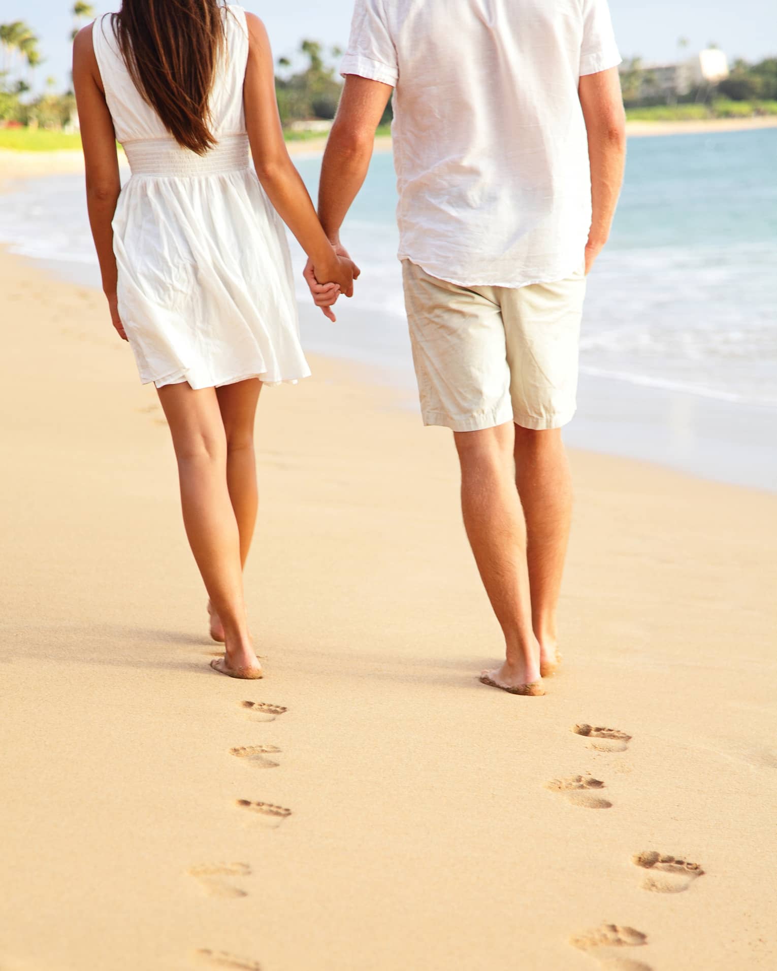 A close-up of a couple holding hands and leaving a trail of footprints as they stroll down a beach near the water’s edge.