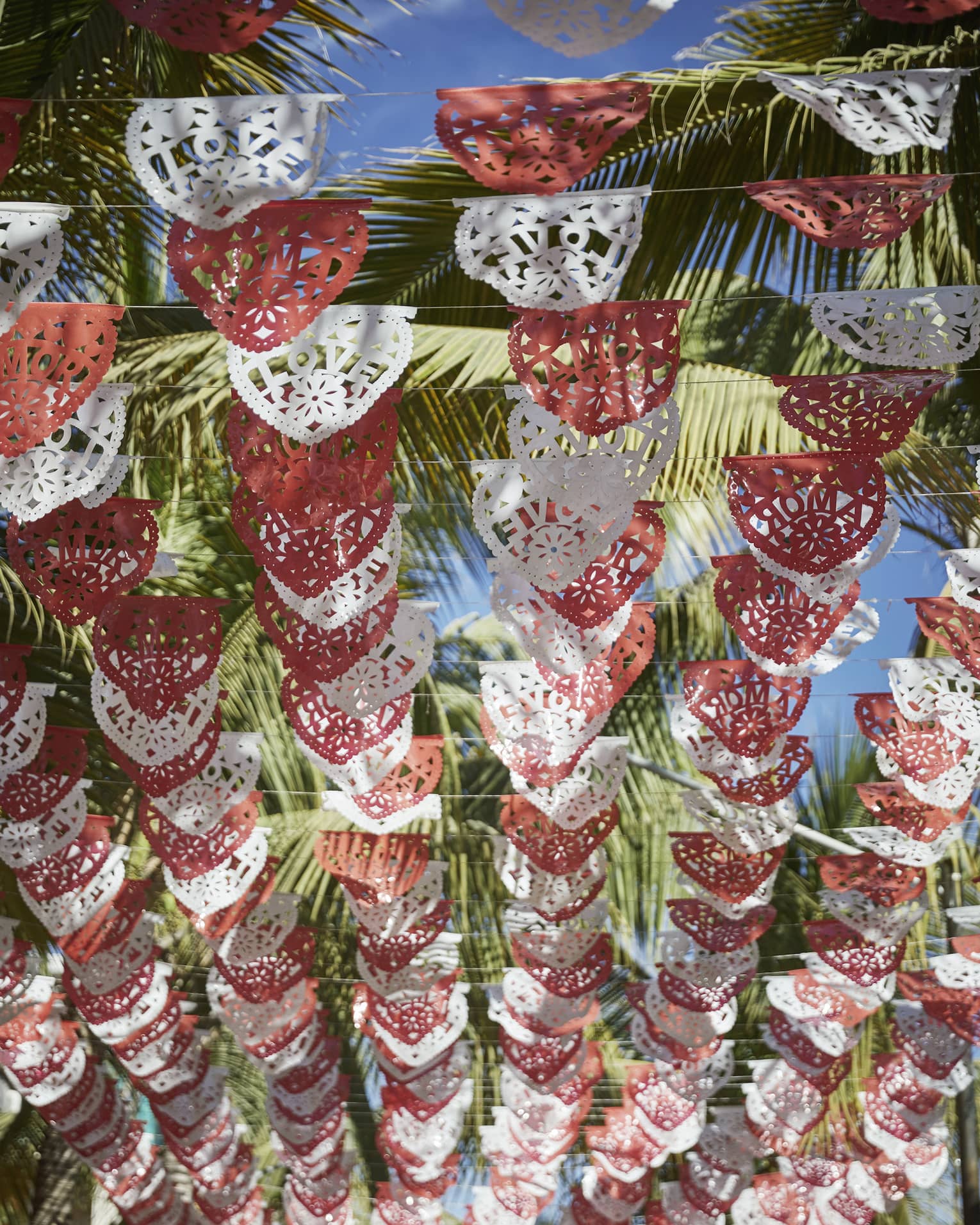 Looking up at rows of red and white Mexican Papel Picado banners hanging above, with palm trees and a sunny sky above them.