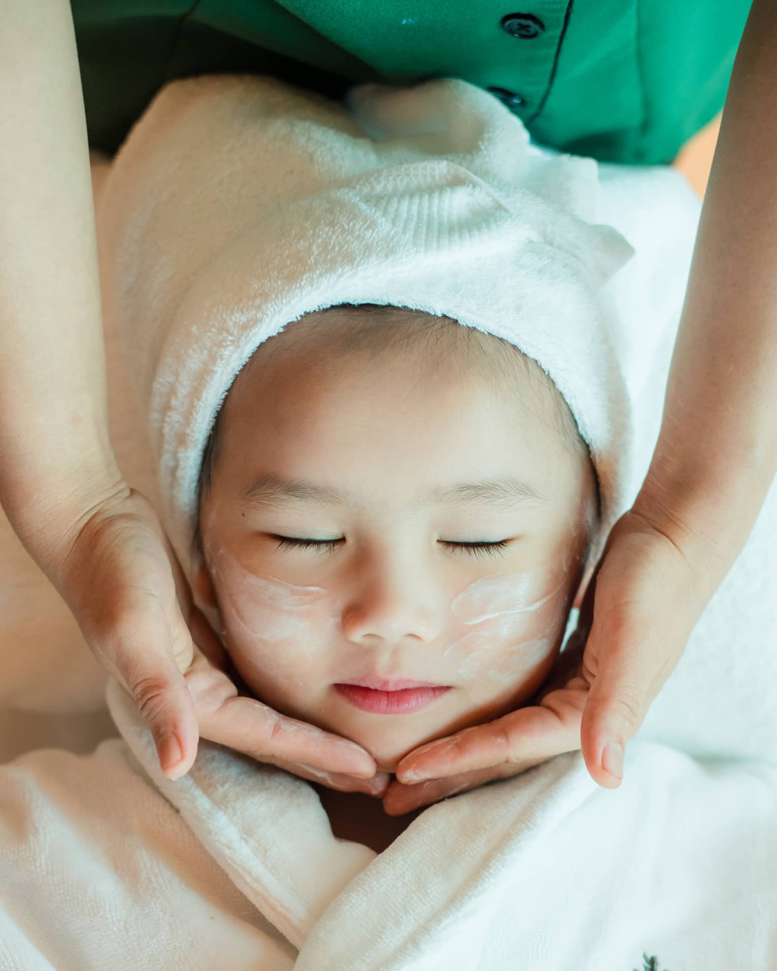 A close-up of a relaxed child wrapped in a towel and robe and enjoying a masseuse massaging a cream into their face.