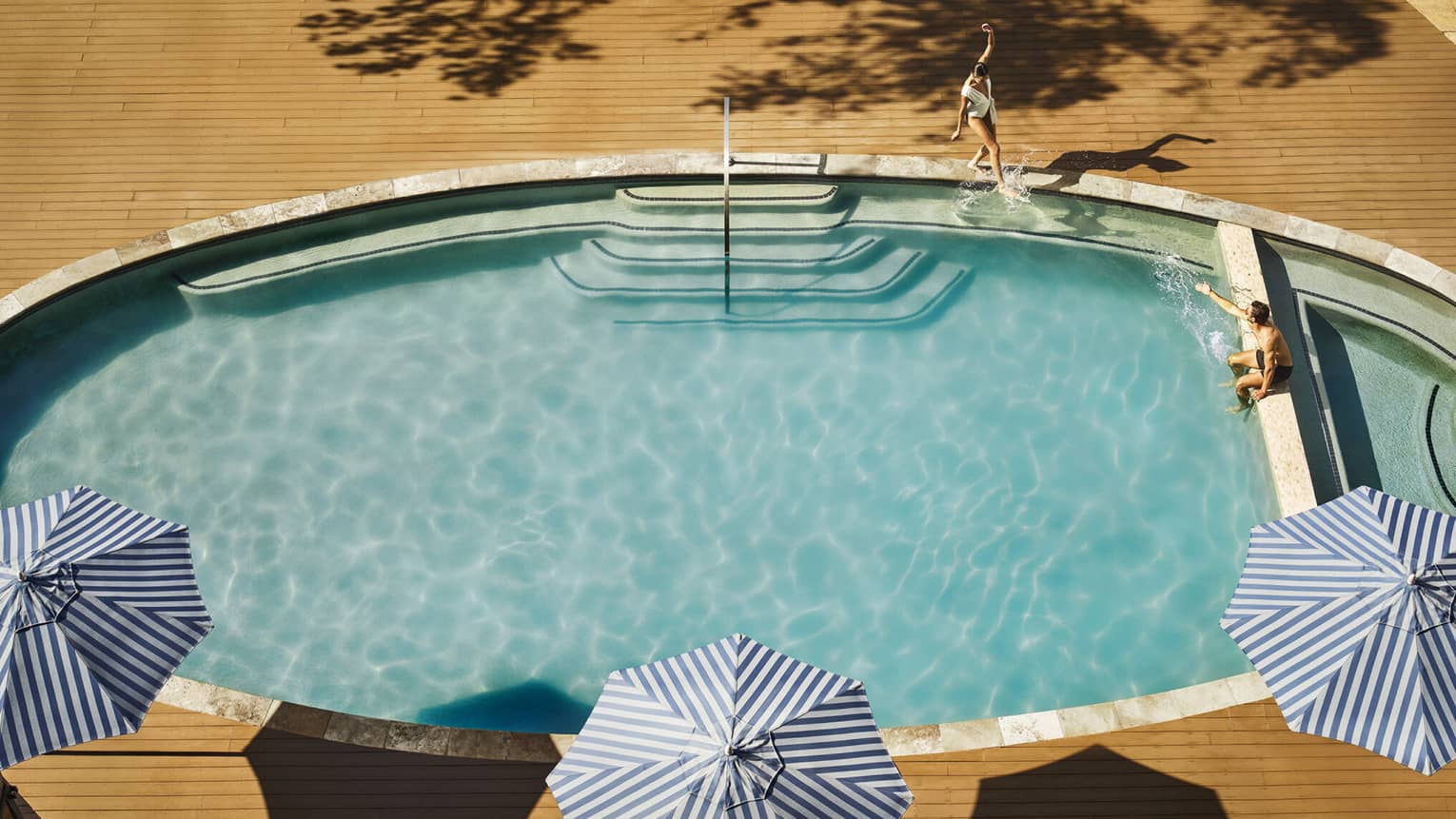 Aerial view of a couple lounging at an oval-shaped pool lined with blue striped umbrellas