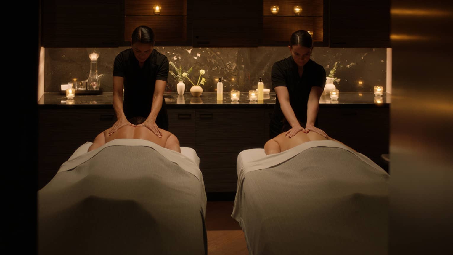 Two people lying face down on massage tables and two massage therapists providing massage in a serene space with low lighting and candles