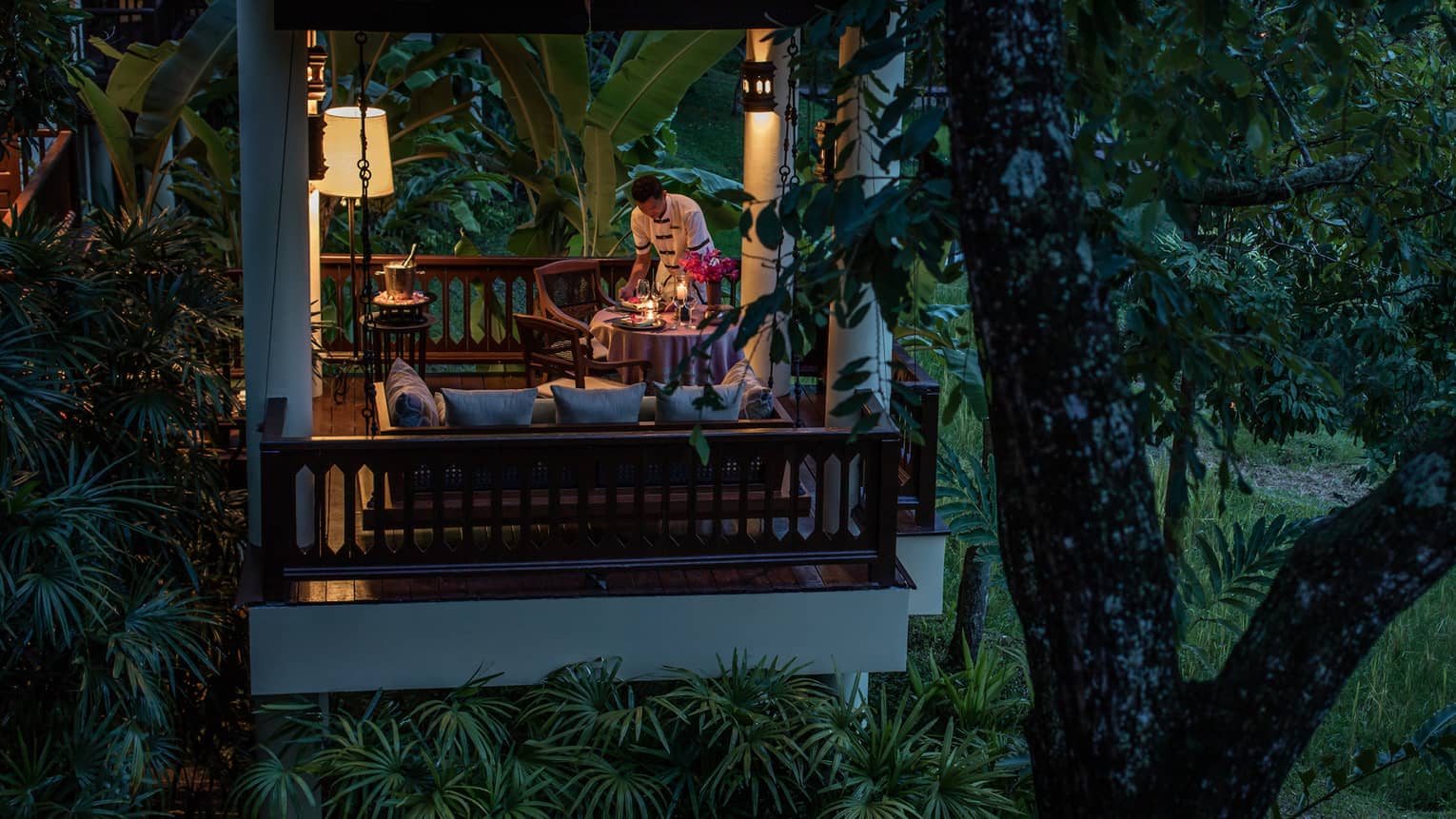 Staff sets candle-lit dining table beneath covered private villa patio at night 