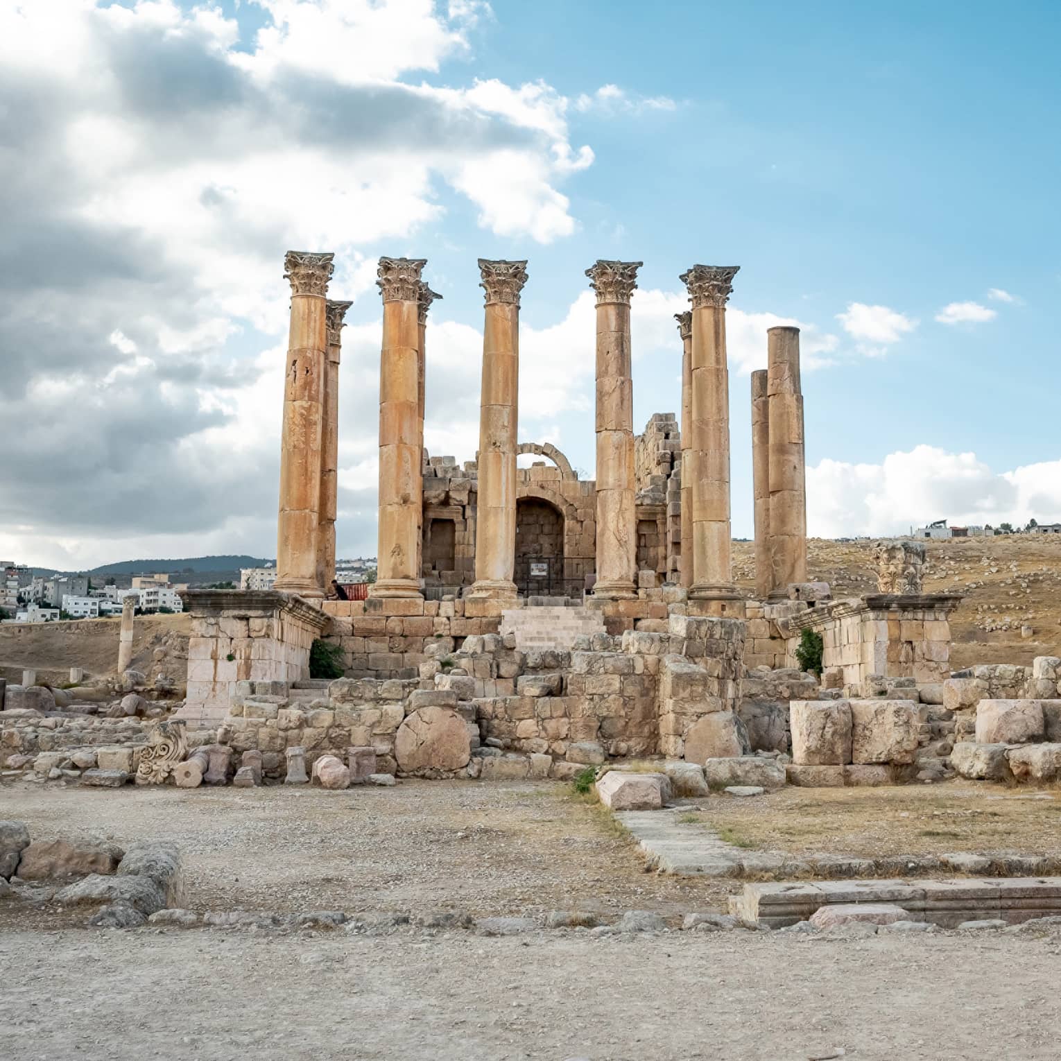 Amongst crumbling ruins, ten ancient Corinthian columns stretch skyward under a cluster of grey and white clouds.