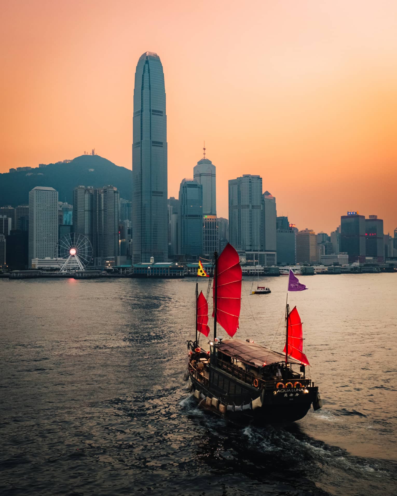 Boat with red sails on harbour in Hong Kong at sunset with city buildings at the shore