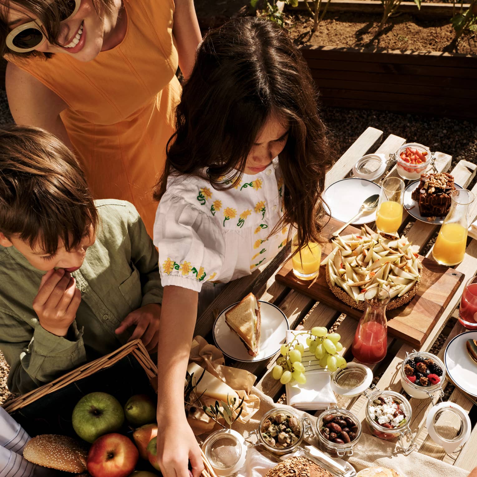 Three people stand over an outdoor picnic table set with a variety of food items, on a bright, sunny day.