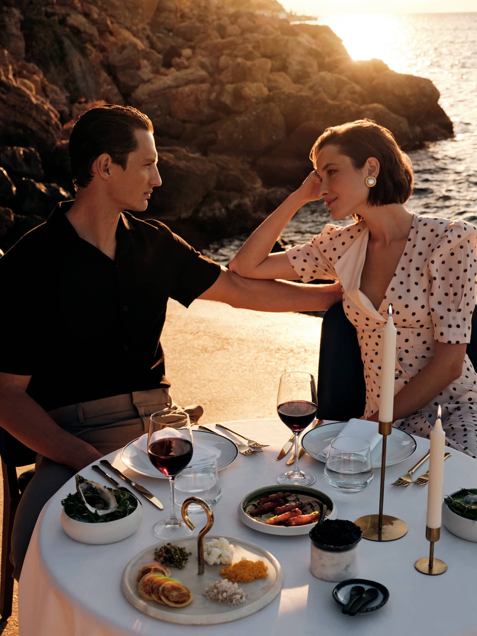 Two people sit at an elegant dining table in an outdoor setting, with a rocky shoreline and the ocean reflecting the sun in the background.