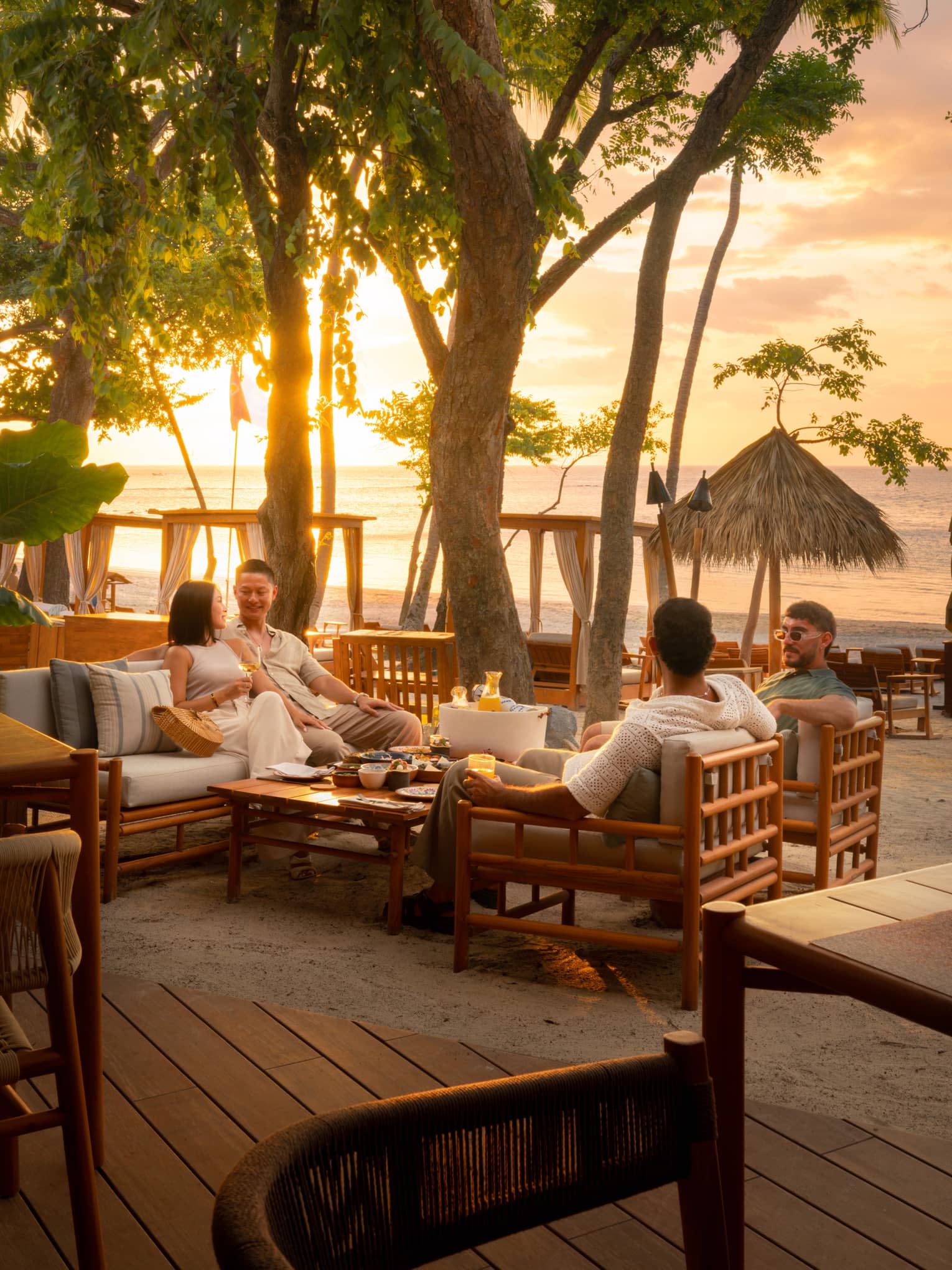 Four people sit smiling and chatting at a beachside dining table as the sun sets behind them