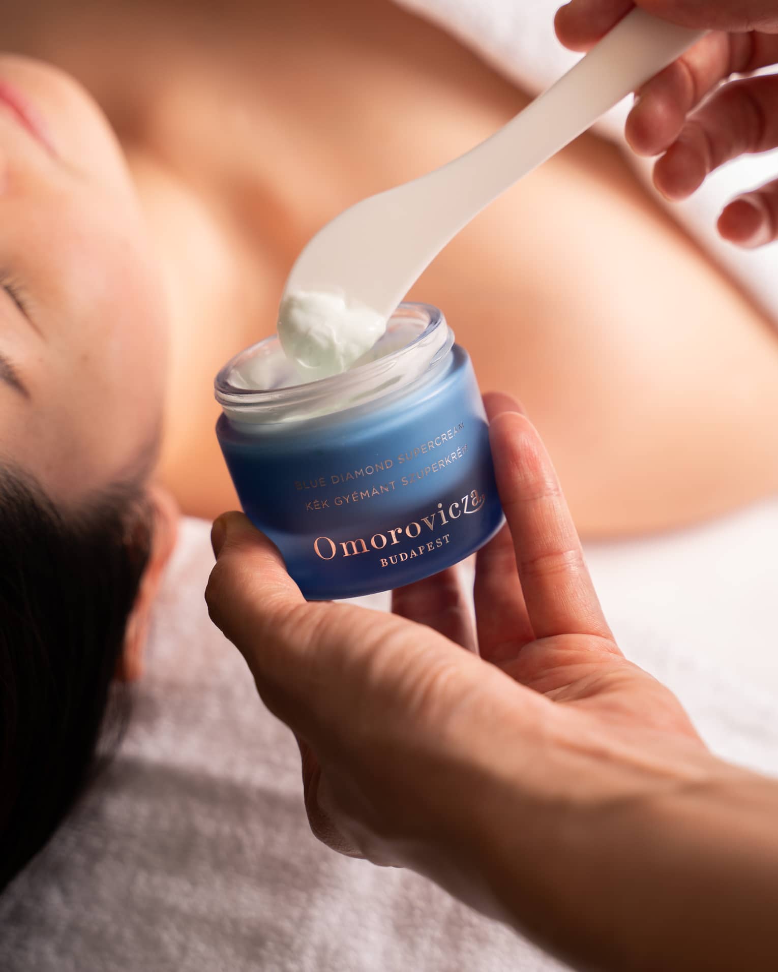 Hand dips spatula into jar of Omorovicza face cream in preparation for woman’s facial