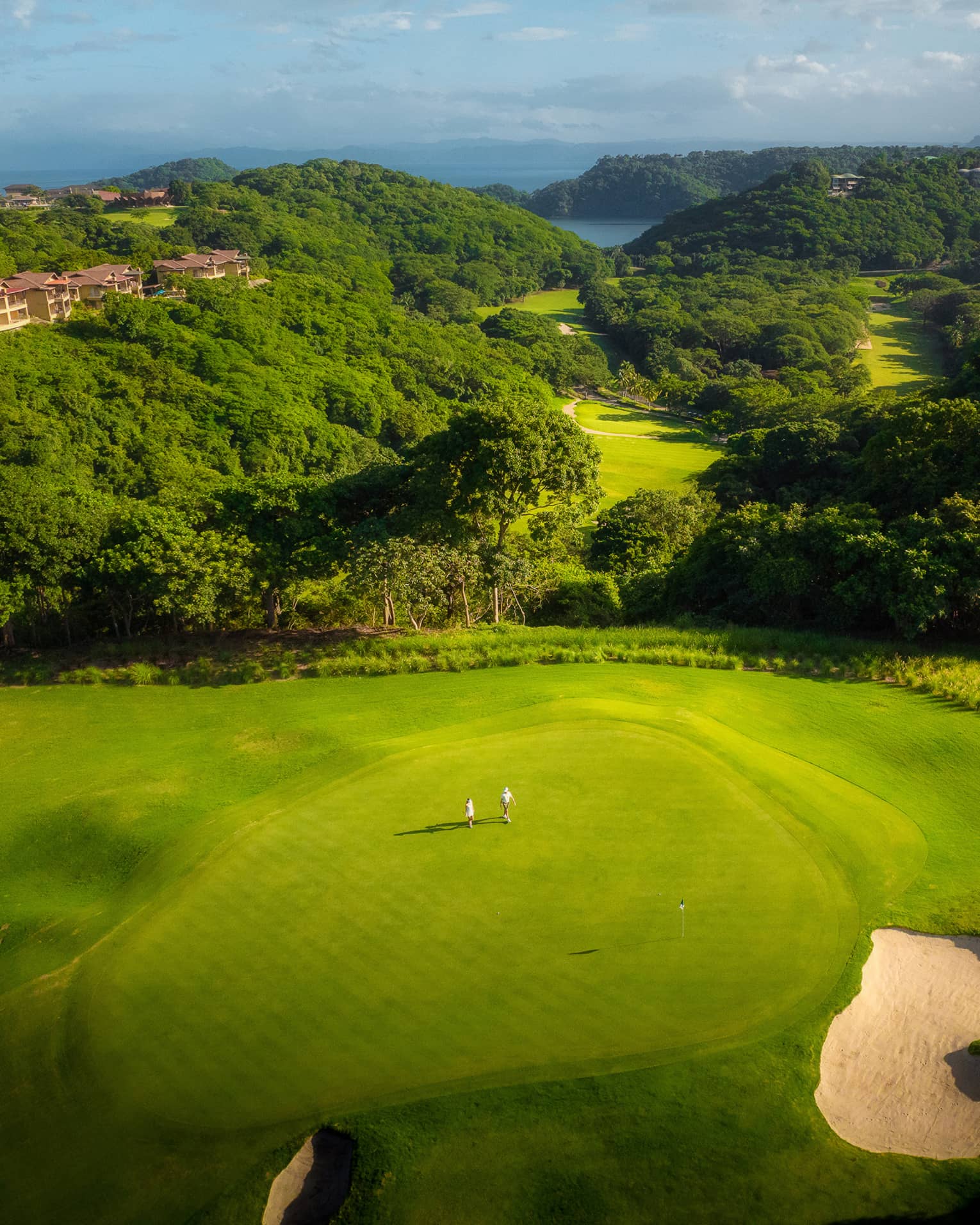 Aerial view of two people standing on a golf green with tree-covered rolling hills behind them leading to the ocean in the distance