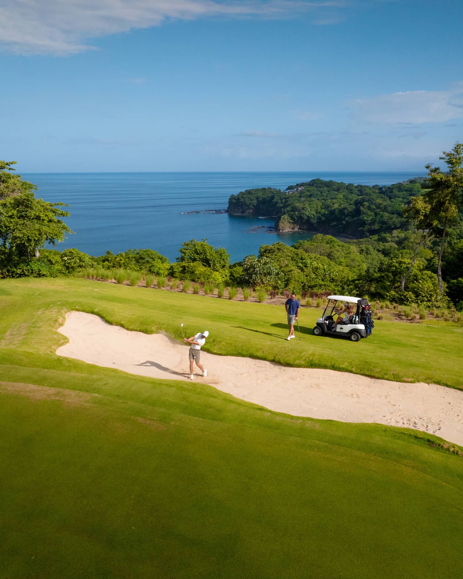 One golfer hits their ball out of a sand trap as another golfer stands by a golf cart looking on, with the ocean in the background