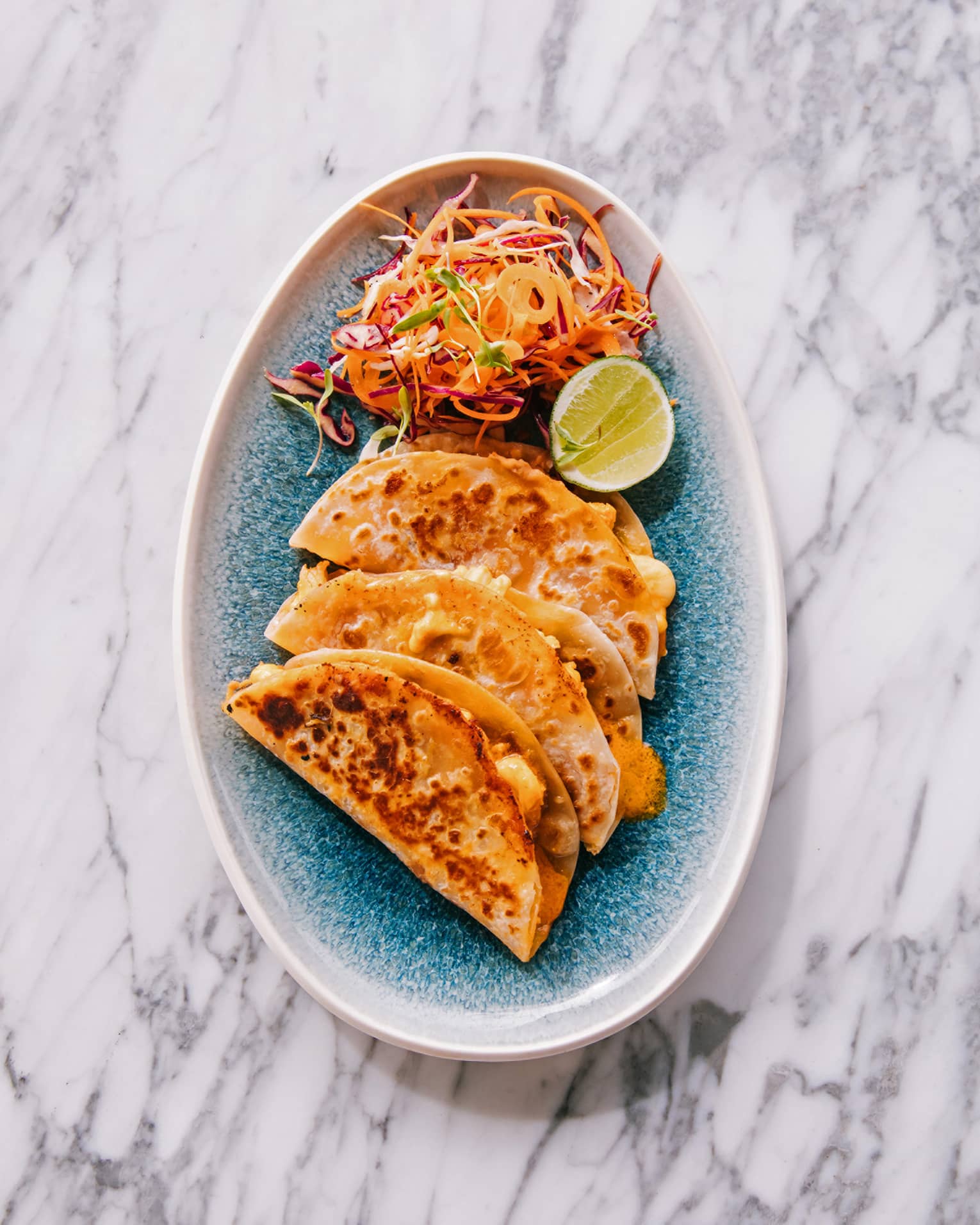 Three tacos and a brightly colored salad with a slice of lime on a blue plate create a visually appealing and appetizing dish.