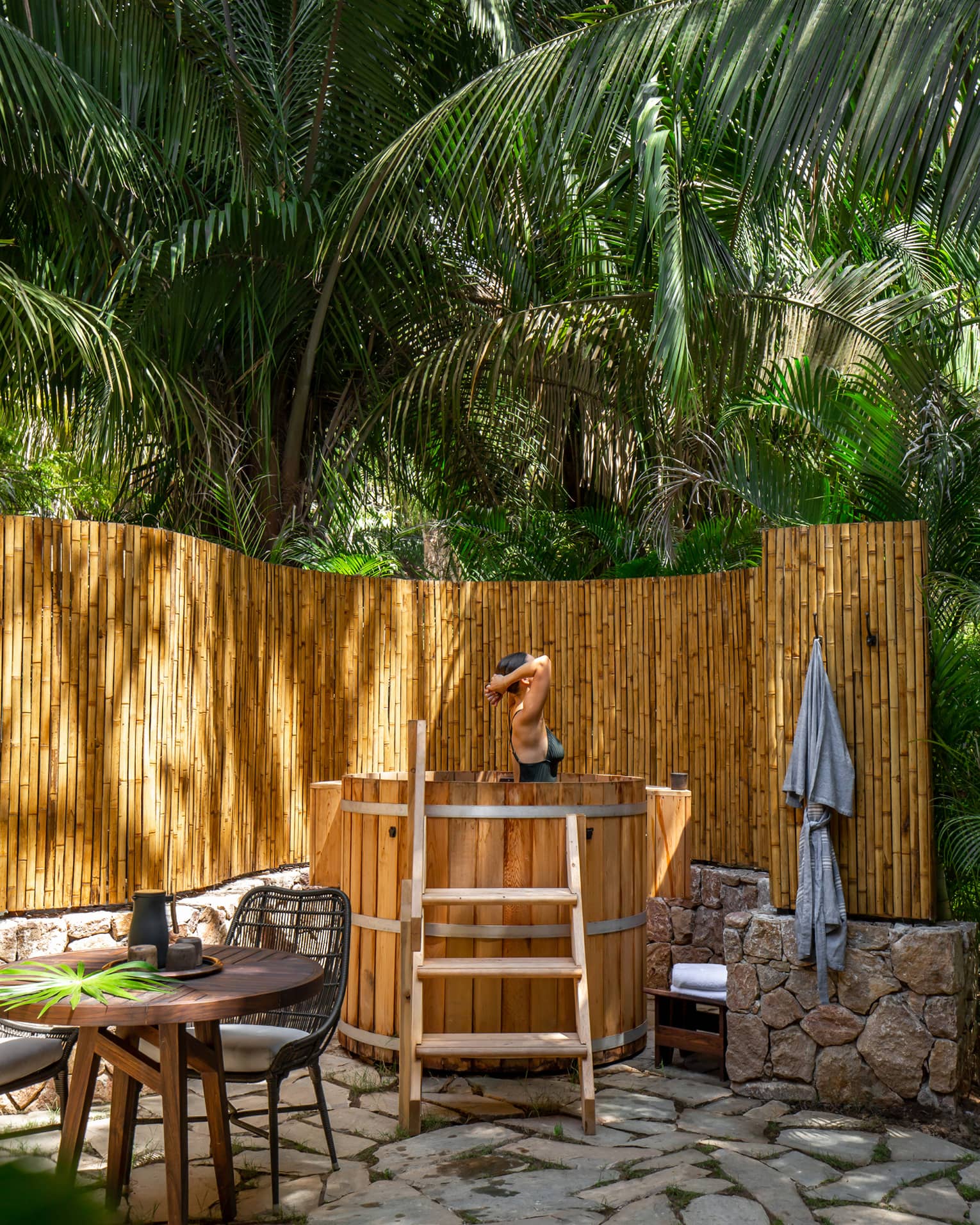 Woman emerges from soak in natural wooden outdoor bath, bamboo fence and palm trees surrounding