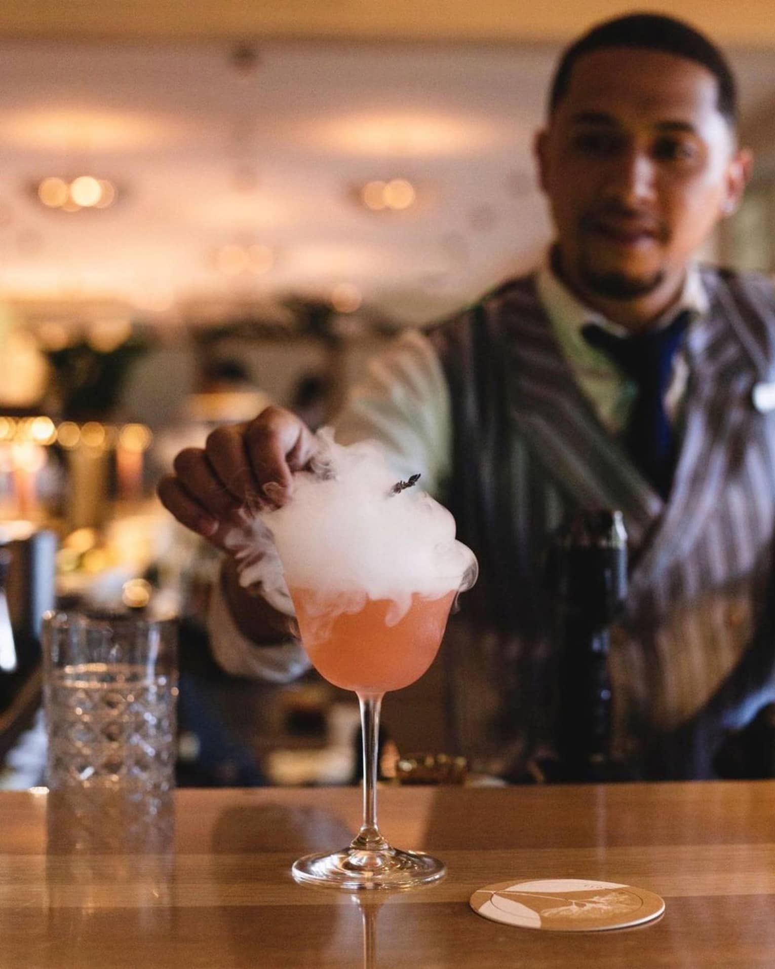A mixologist garnishes a bright orange cocktail atop a smooth wooden bar; smoke billows over the sides of the stemmed glass.