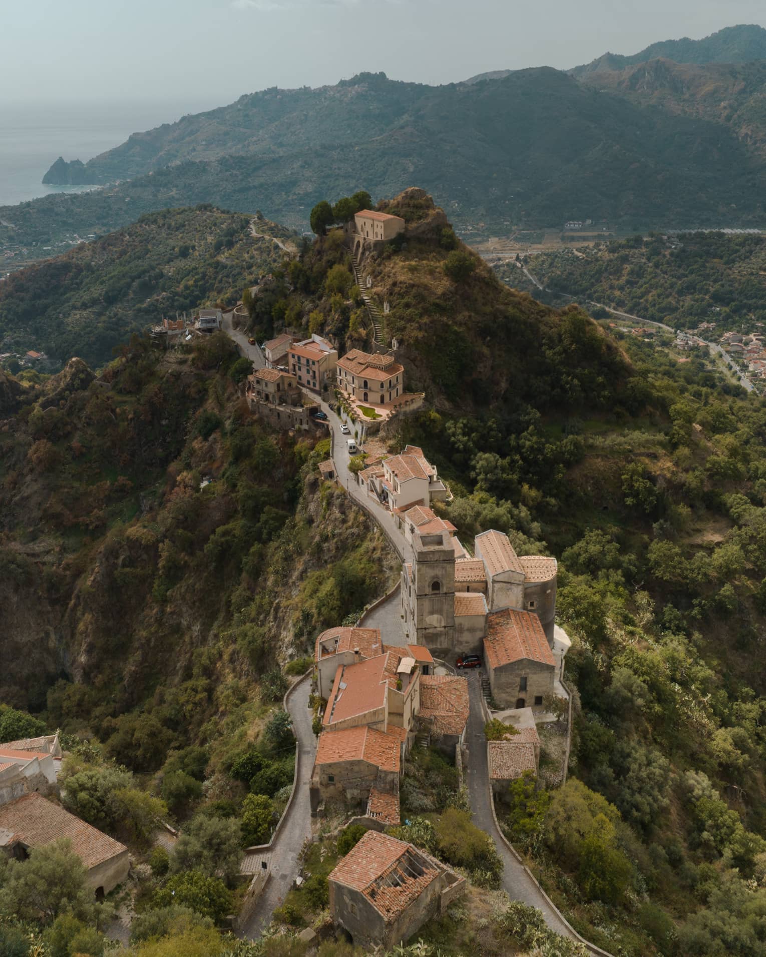 Aerial view of a historic hilltop village with winding roads, stone buildings and terracotta roofs, nestled in a lush green mountain landscape.